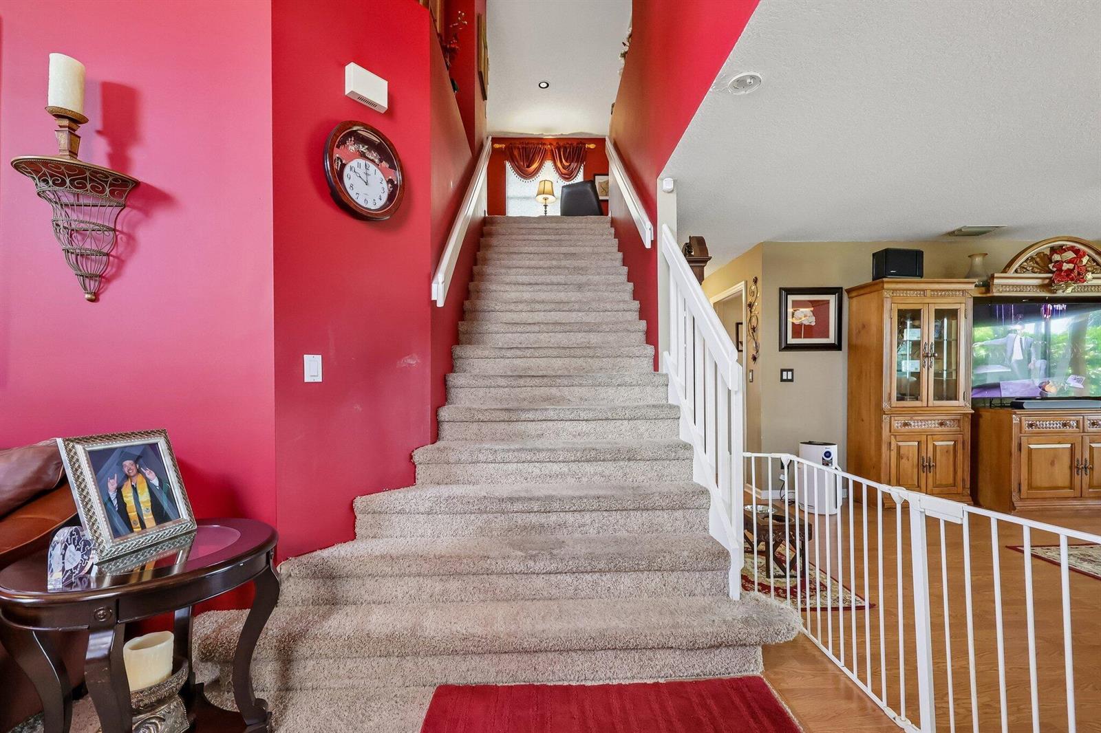 A carpeted staircase with white railings ascends between a red wall and a living area — 6205 Lansdowne Circle, Boynton Beach FL 33472, BOYNTON BEACH, FL