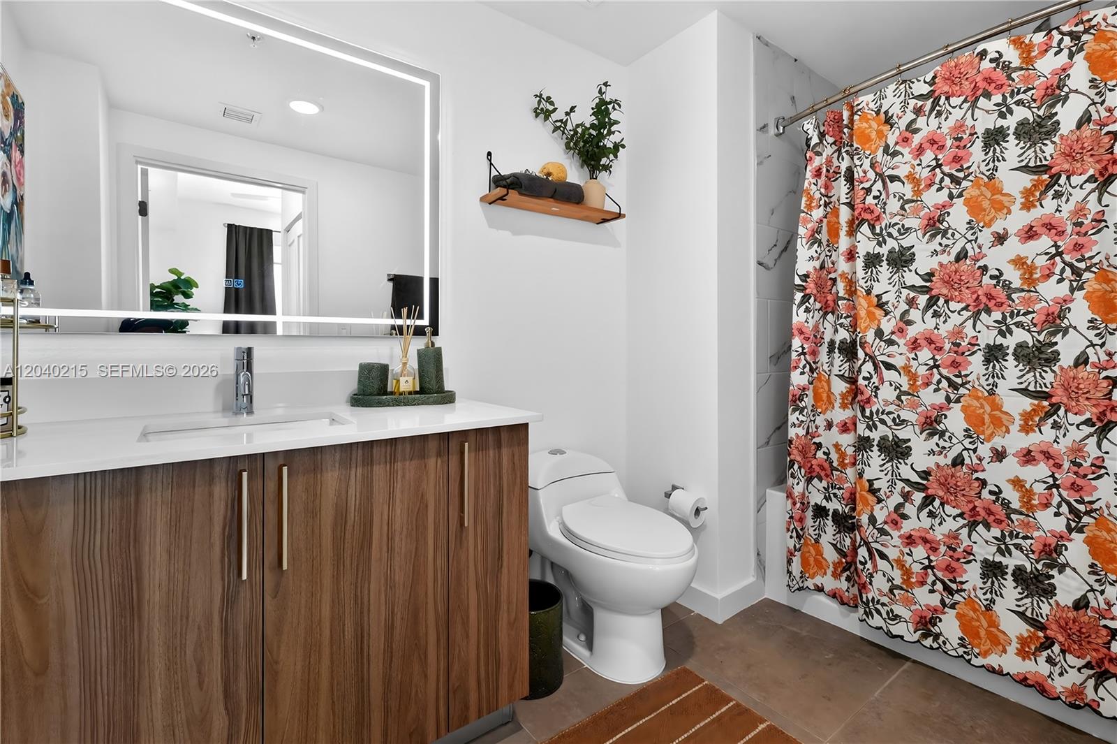 A modern bathroom features a wood vanity, a backlit mirror, and a floral shower curtain — 2001 Van Buren St # 504, Hollywood FL 33020, HOLLYWOOD, FL