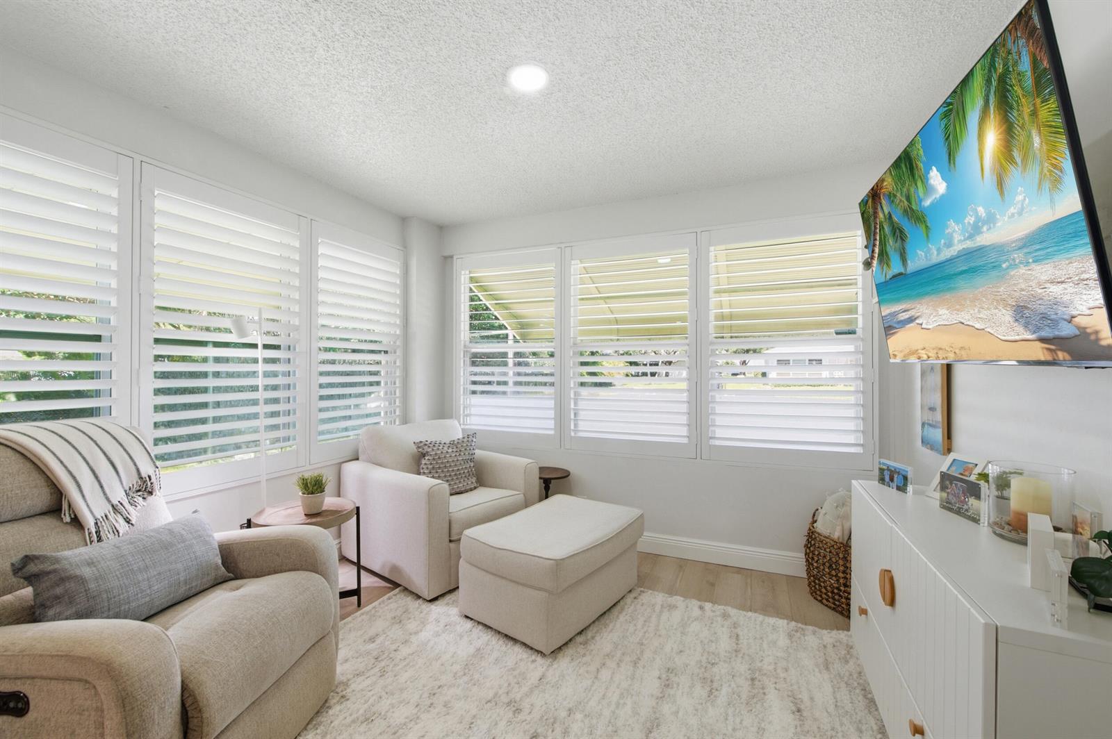 A bright living room features white plantation shutters, a television, and two comfortable armchairs — 957 Circle Drive # D, Delray Beach FL 33445, DELRAY BEACH, FL