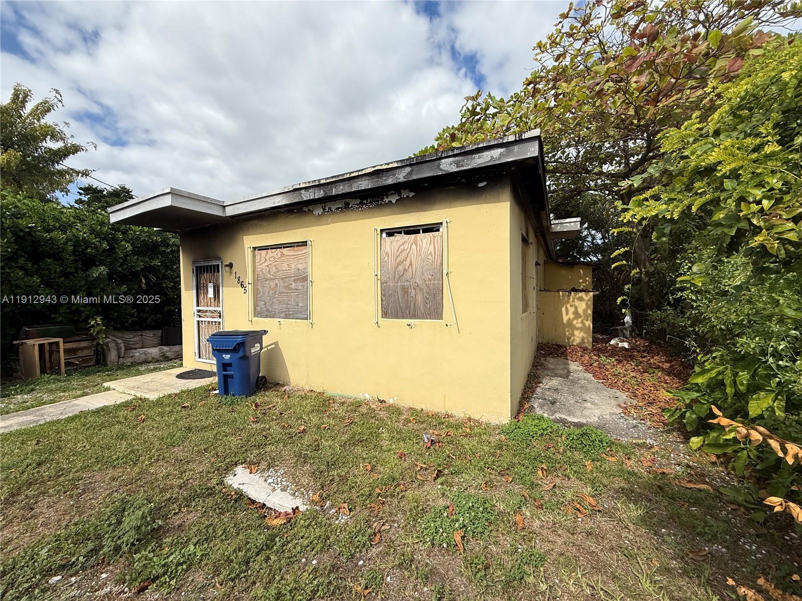 A small, yellow, single-story house with boarded-up windows and a blue trash bin outside — 1865 NW 70th St, Miami FL 33147, MIAMI, FL