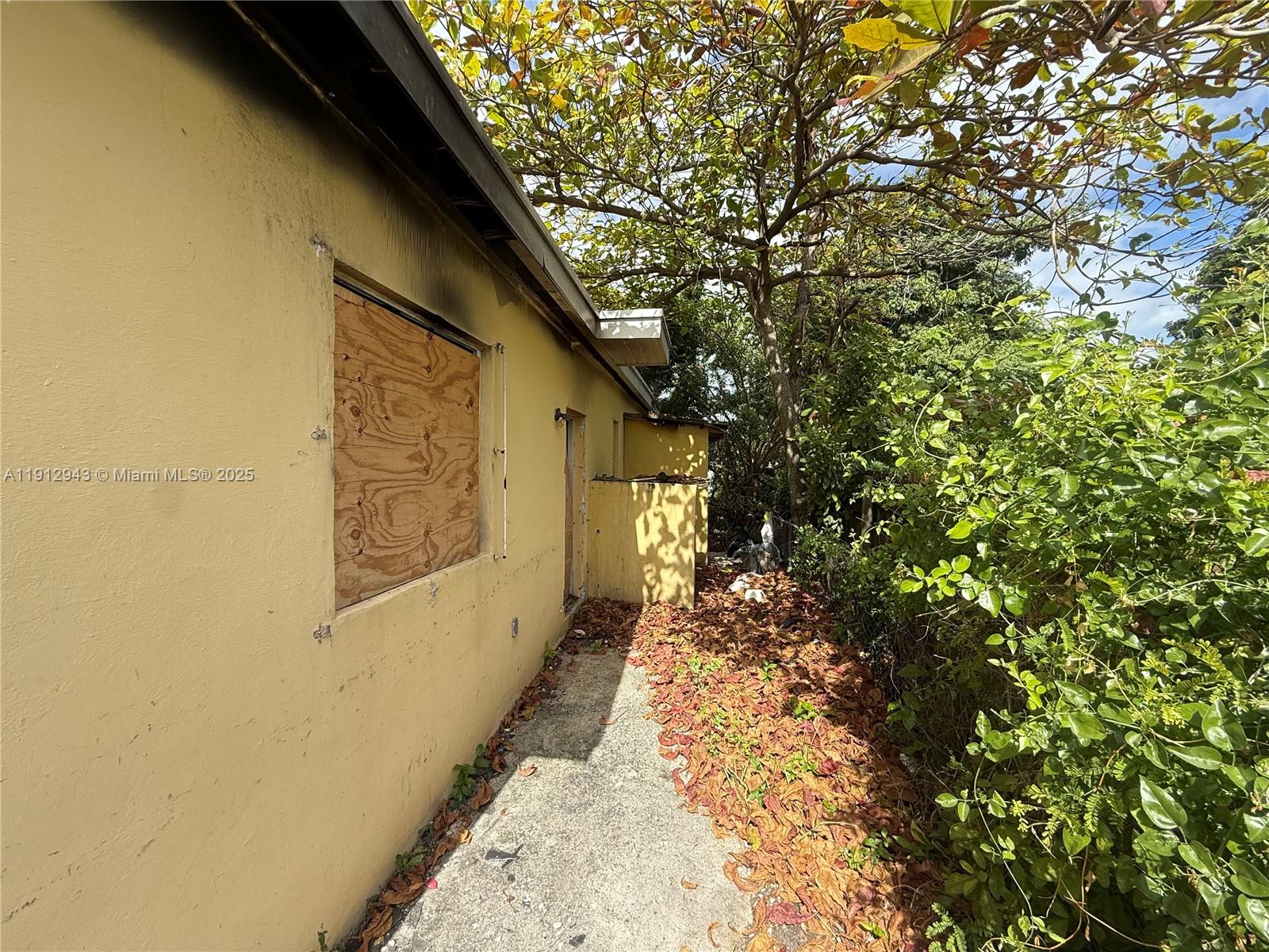 The exterior of a yellow house features a boarded-up window and a concrete walkway — 1865 NW 70th St, Miami FL 33147, MIAMI, FL
