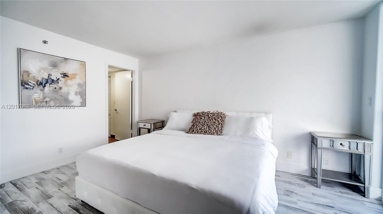 A bedroom features a white bed, two mirrored nightstands, and a large abstract wall painting — MIAMI BEACH, FL