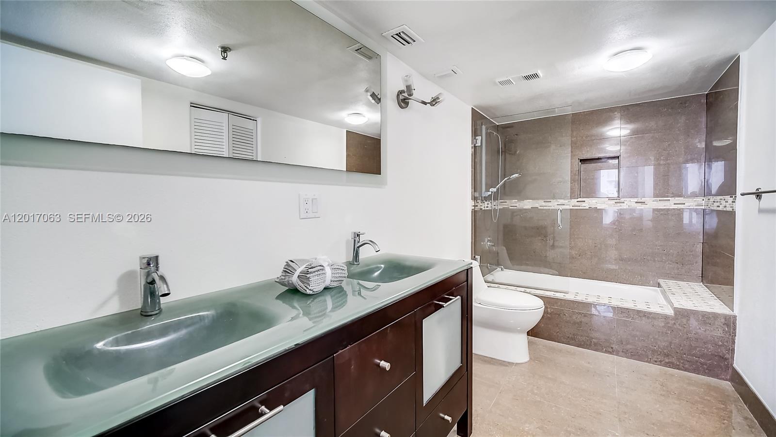 A bathroom features a double vanity with a glass countertop and a tiled shower-tub combination — MIAMI BEACH, FL