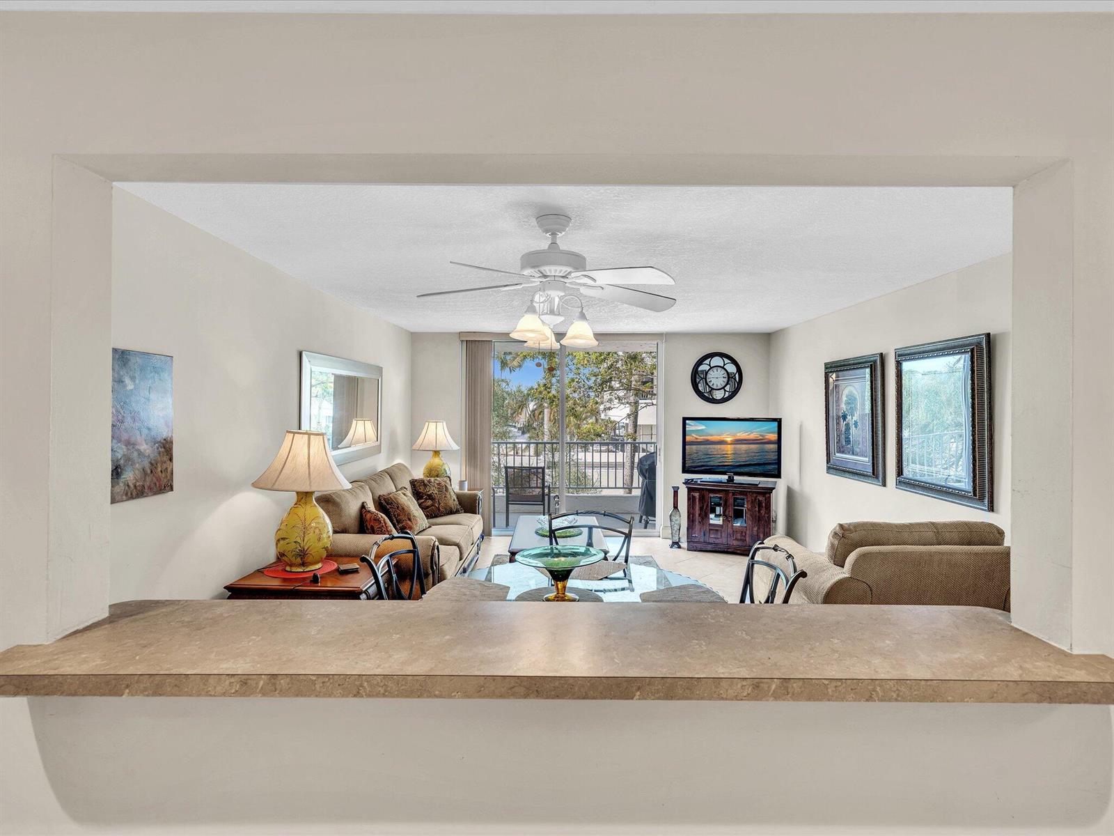 A view through a kitchen pass-through window into a living room with a balcony — 1421 S Ocean Boulevard # 219, Pompano Beach FL 33062, POMPANO BEACH, FL