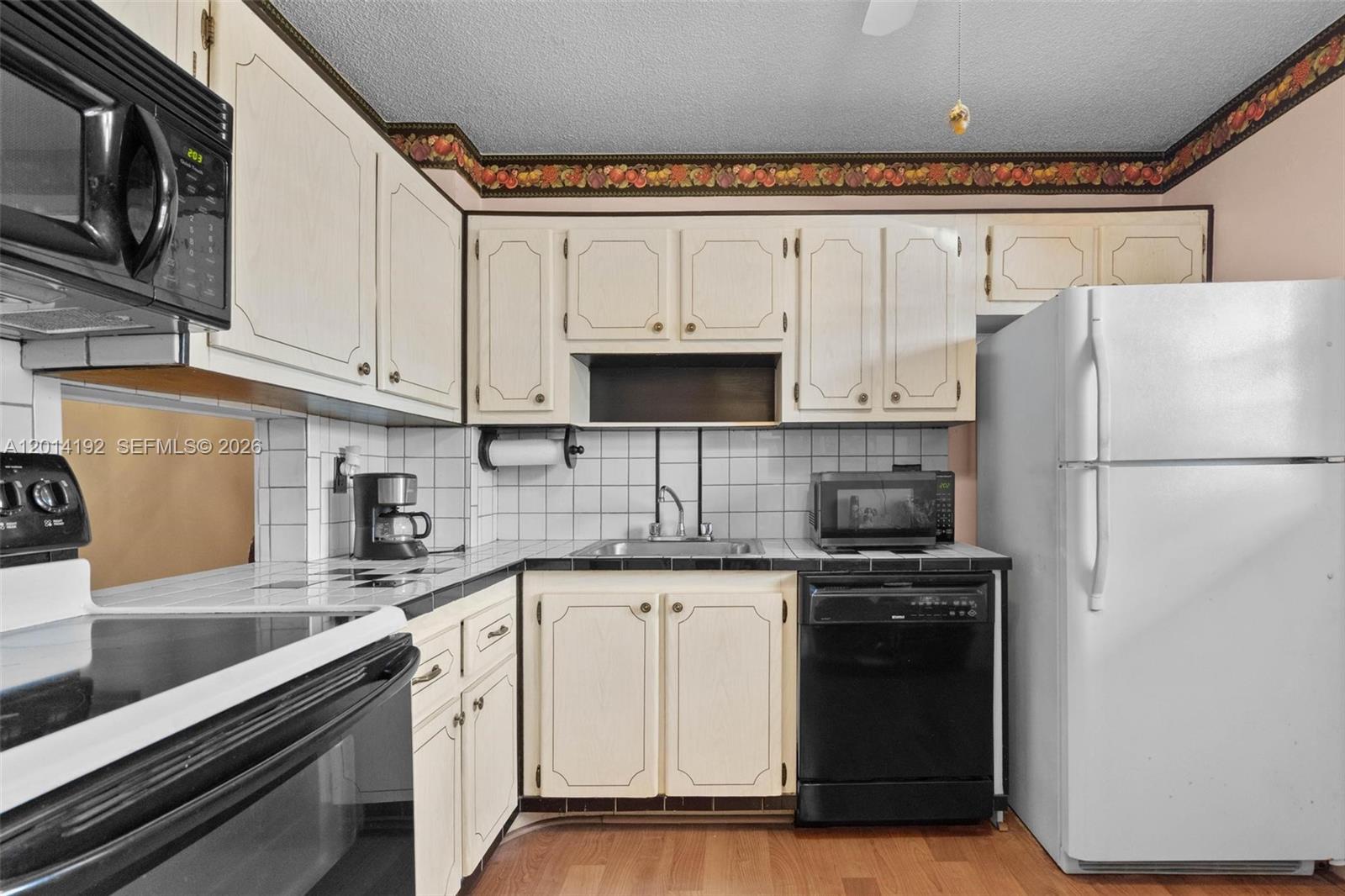 A kitchen features white cabinets, black appliances, tiled countertops, and a fruit-patterned wallpaper border — 100 Berkley Rd # 106, Hollywood FL 33024, HOLLYWOOD, FL
