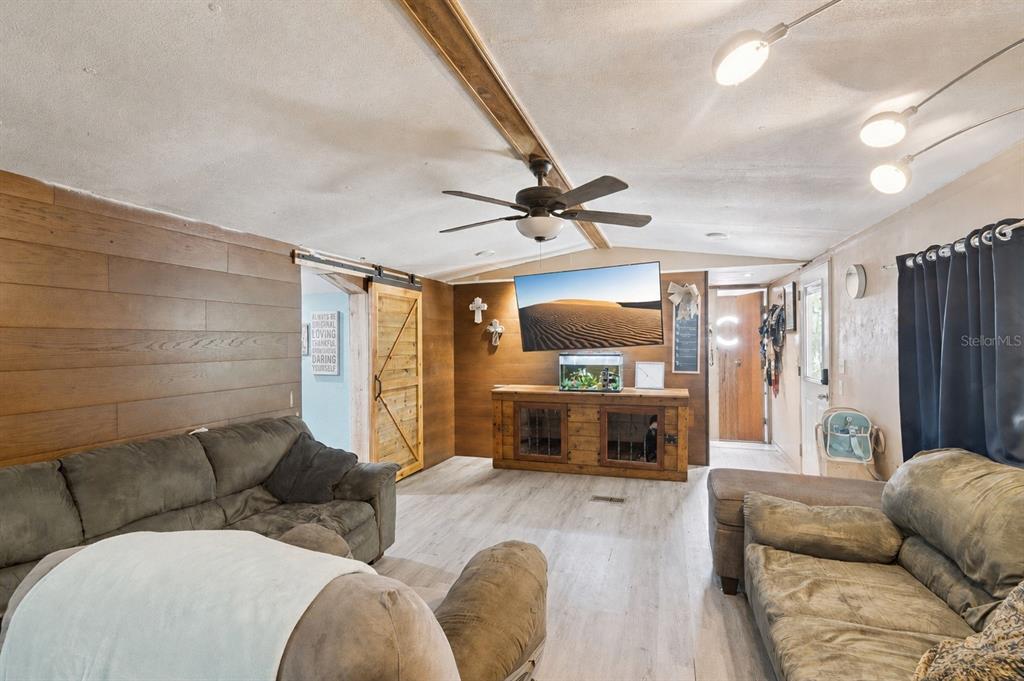 A living room with wood-paneled walls, a sliding barn door, and light-colored wood-look flooring — 10927 LUANA DRIVE, LEESBURG, FL