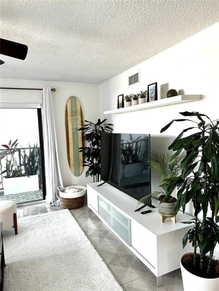 A living room features a television on a white console, a surfboard, and potted plants — 1975 SE 3rd Street # 403, Deerfield Beach FL 33441, DEERFIELD BEACH, FL