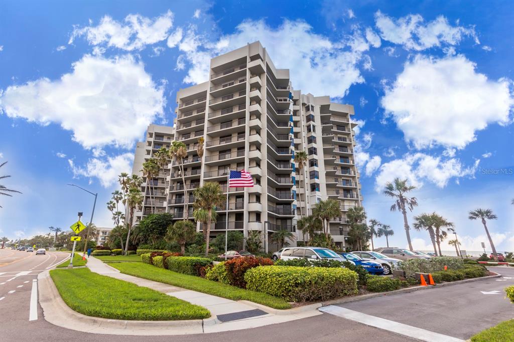 A tall, multi-story residential building with balconies stands behind a landscaped parking area and road — 1600 GULF BOULEVARD UNIT 414, CLEARWATER, FL