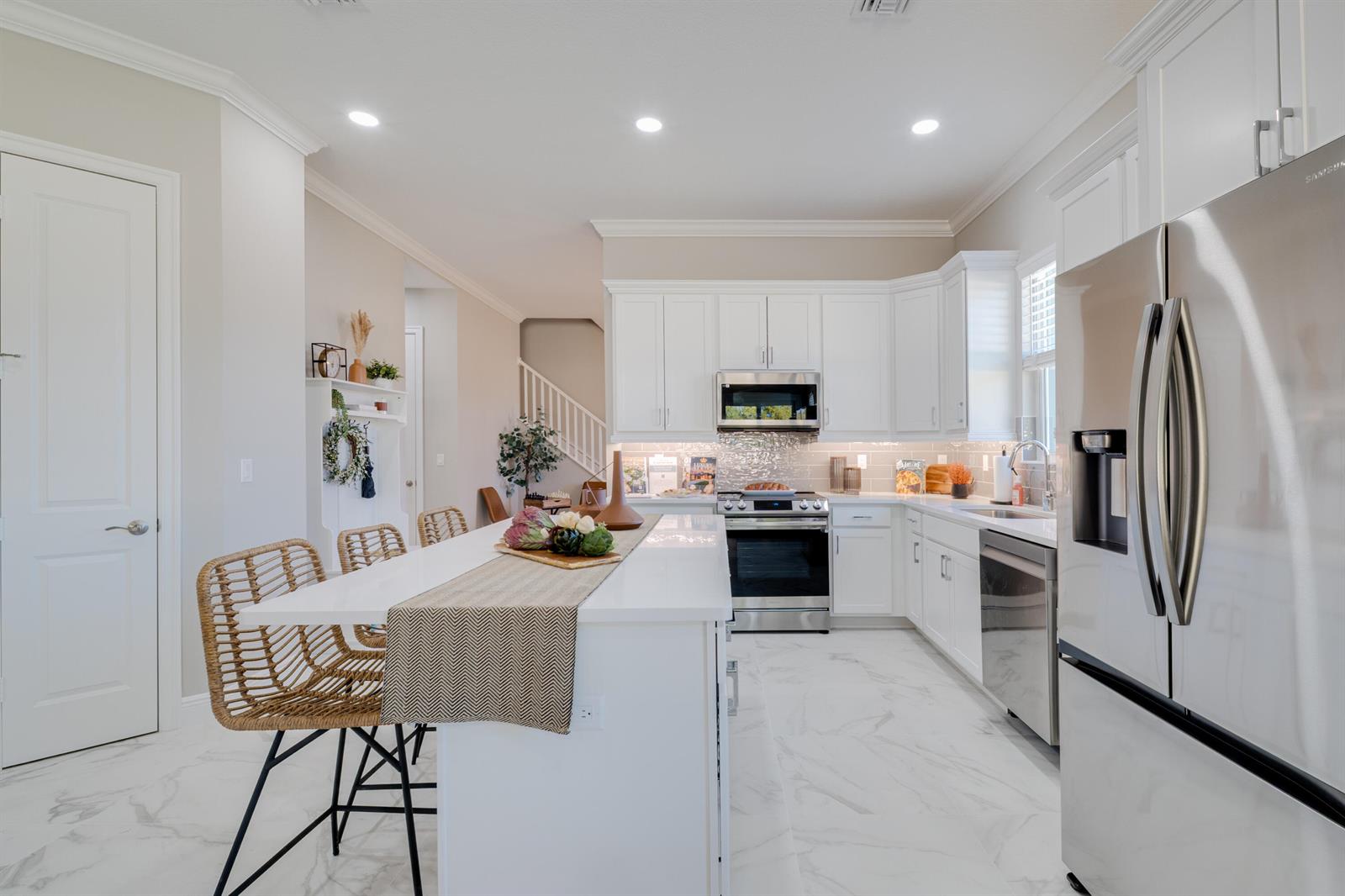 A modern white kitchen features a marble-topped island, stainless steel appliances, and woven bar stools — 2005 Bridgepointe Circle # 97, Vero Beach FL 32967, VERO BEACH, FL