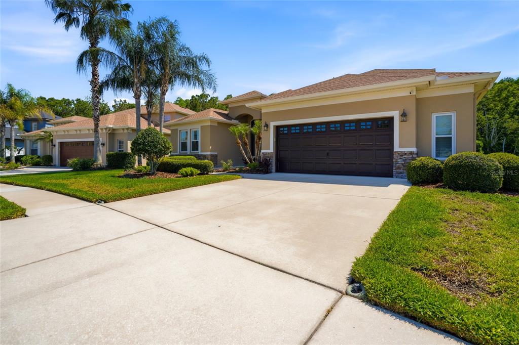 A single-story suburban house features a tan stucco exterior, a dark garage door, and landscaping — 3320 GRASSGLEN PLACE, WESLEY CHAPEL, FL