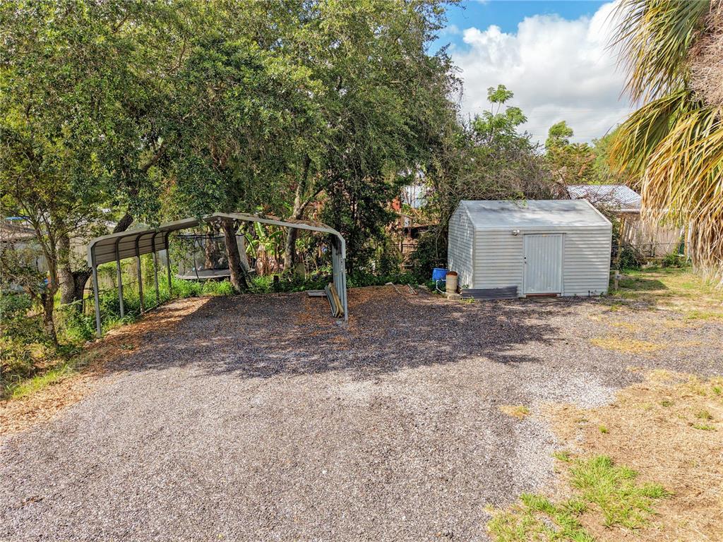 A gravel driveway features a metal carport and a small storage shed near mature trees — 120 CHESNEY BOULEVARD, FROSTPROOF, FL
