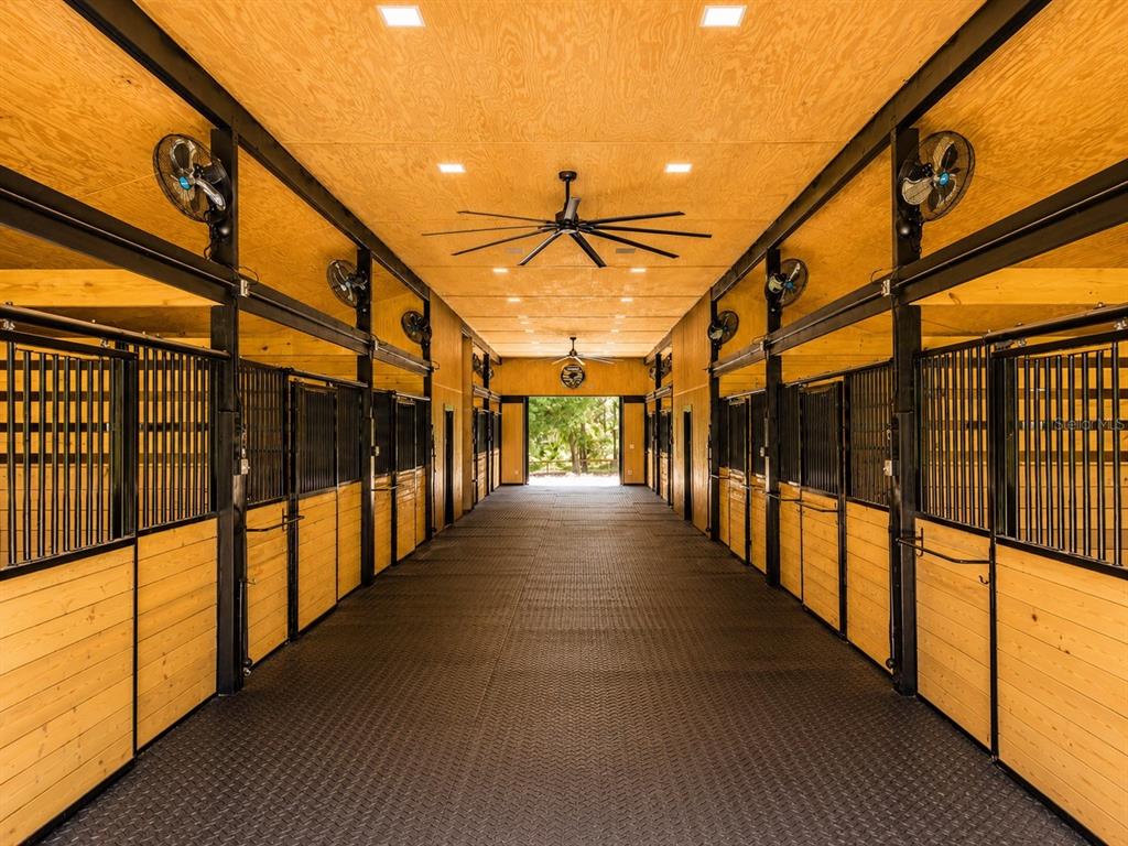 A clean, modern horse stable aisle with wood-paneled walls, black metal stalls, and ceiling fans — 196 NW 100TH AVENUE, OCALA, FL