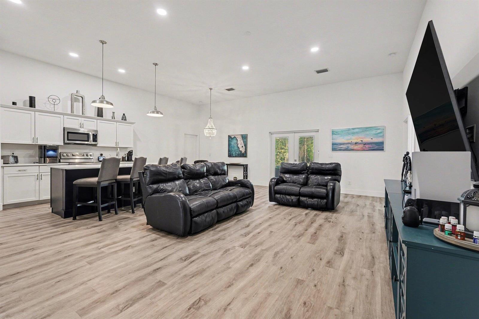 A spacious open-concept living room and kitchen featuring white cabinetry, black leather furniture, and wood-look flooring — 1444 Seeley Circle NW, Palm Bay FL 32907, PALM BAY, FL