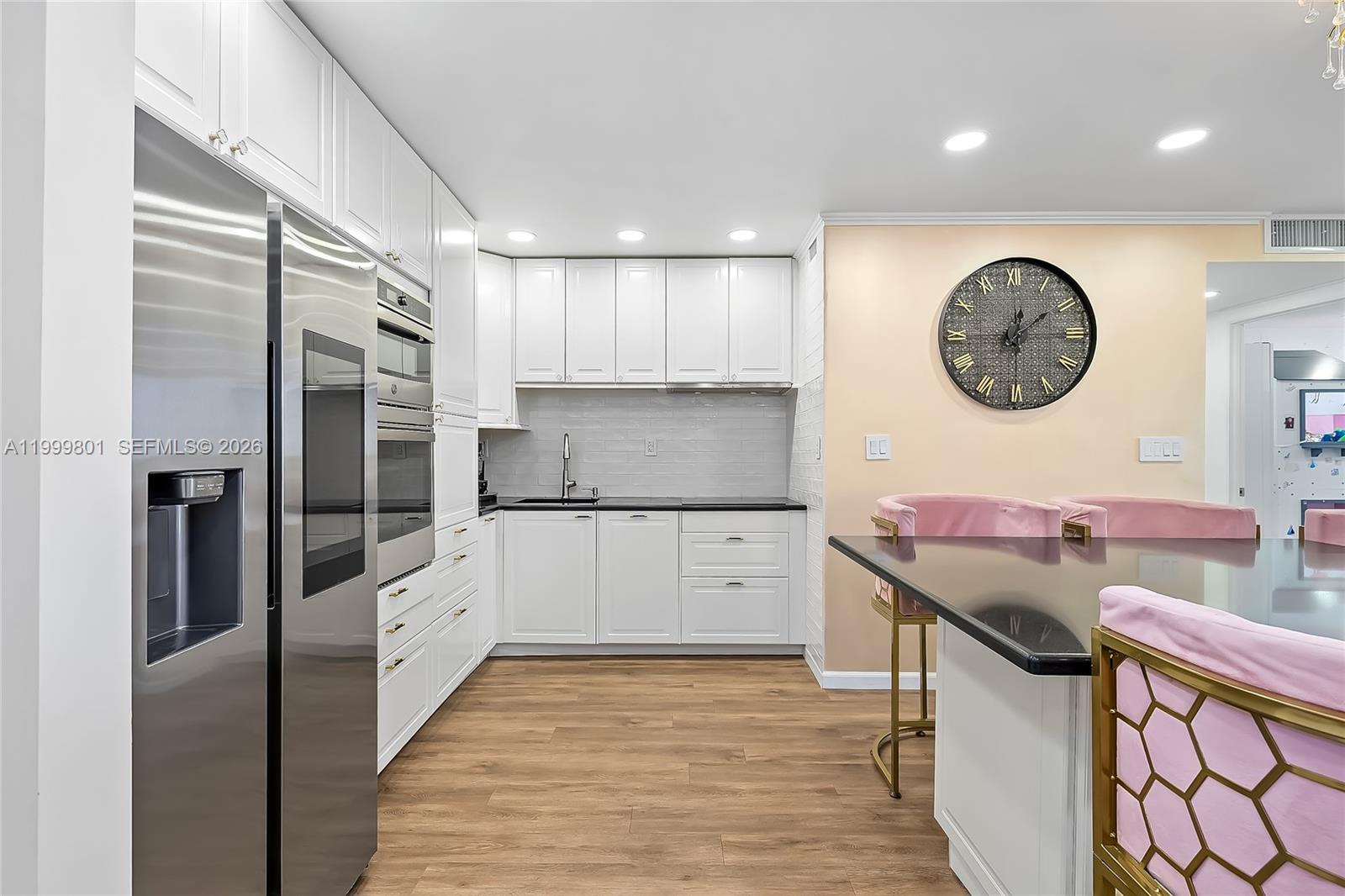 A modern kitchen features white cabinets, stainless steel appliances, black countertops, and pink bar stools — 600 Three Islands Blvd # 604, Hallandale Beach FL 33009, HALLANDALE BEACH, FL