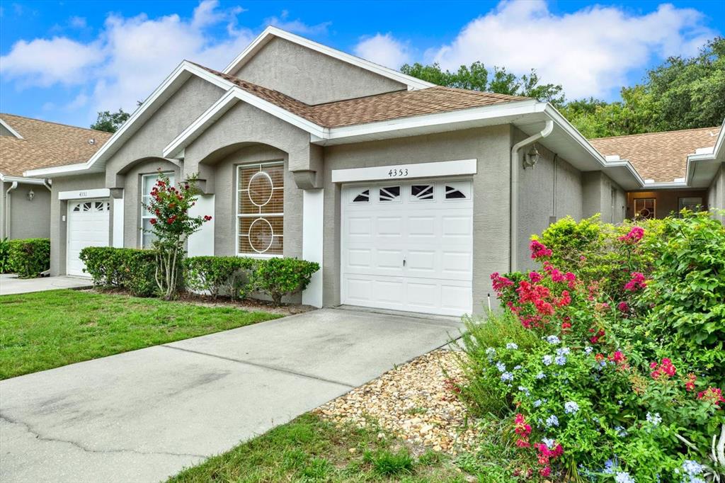 A beige single-story house with a white garage door, brown roof, and landscaped front yard — 4353 BROOKER CREEK DR, PALM HARBOR, FL