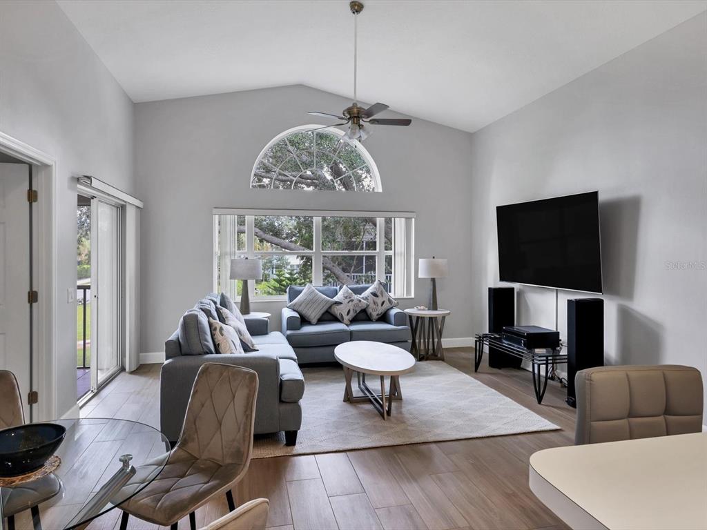 A living room with vaulted ceilings, a large arched window, gray sofas, and wood flooring — 7234 ELEANOR CIRCLE UNIT 203, SARASOTA, FL
