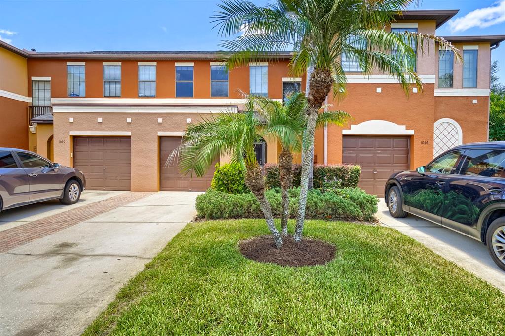 A two-story townhouse exterior features a palm tree, a small lawn, and two garage doors — 1048 TULLAMORE DRIVE, WESLEY CHAPEL, FL