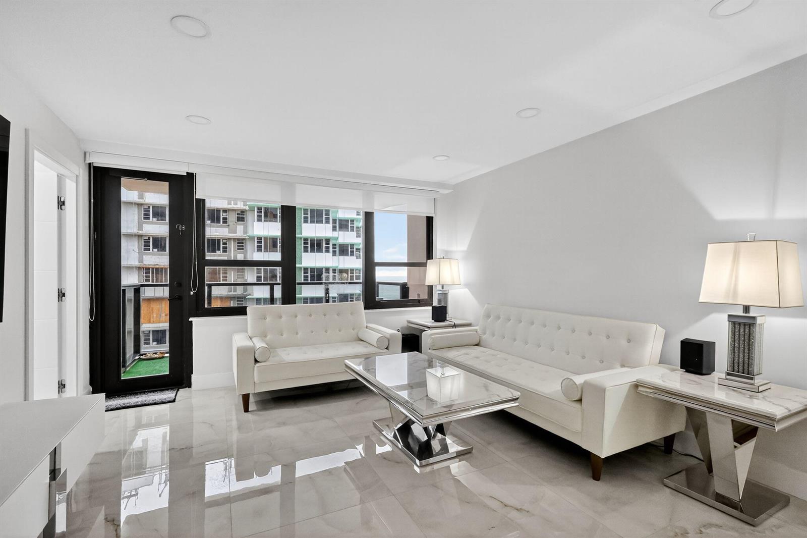 A bright living room features white tufted sofas, a glass coffee table, and marble flooring — 5225 Collins Avenue # 1408, Miami Beach FL 33140, MIAMI BEACH, FL