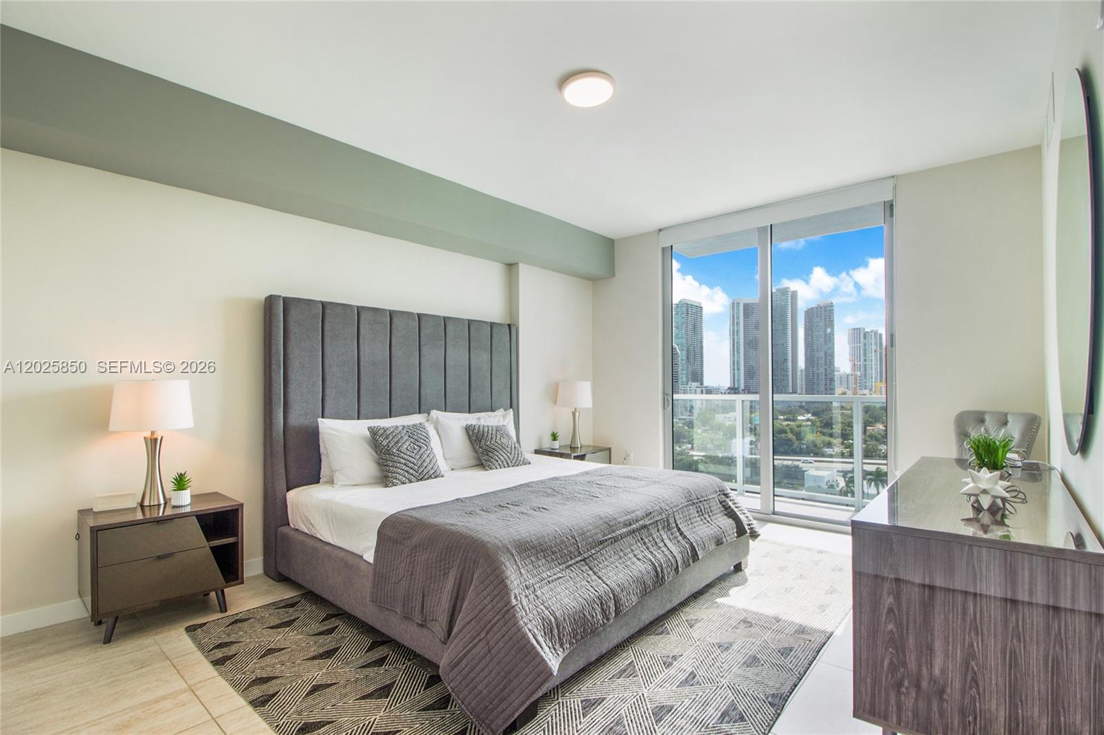 A modern bedroom features a gray upholstered bed, geometric rug, and floor-to-ceiling city view windows — 3900 Biscayne Blvd # 1110, Miami FL 33137, MIAMI, FL