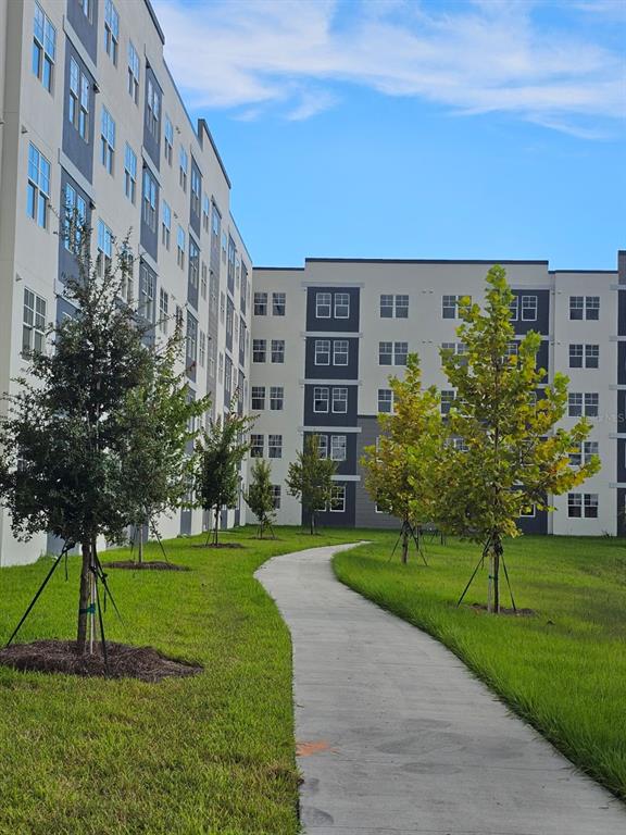 A concrete path winds through a grassy lawn between two modern apartment buildings with trees — 5587 MILLENIA PARK BOULEVARD UNIT 402, ORLANDO, FL