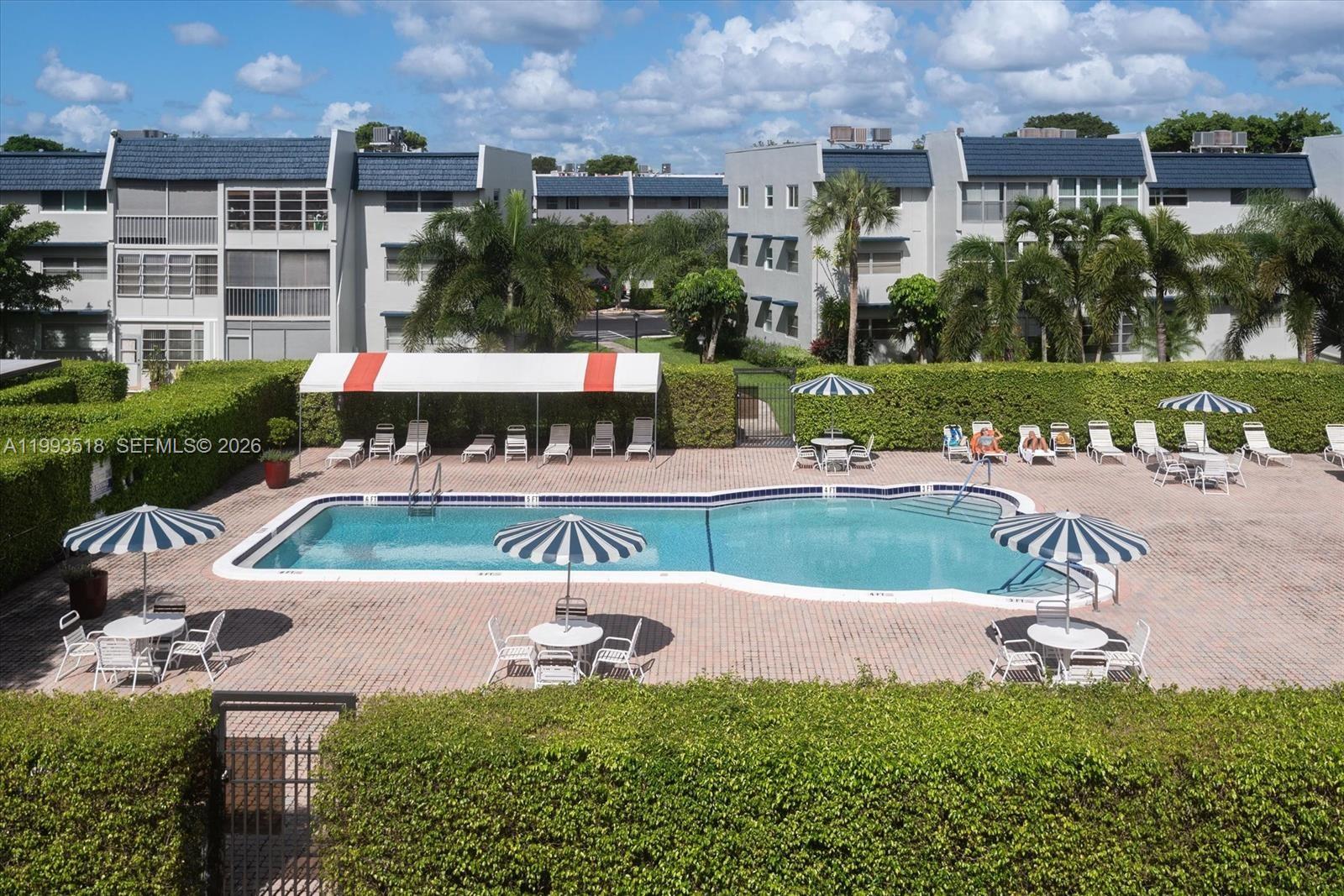 An outdoor swimming pool surrounded by a brick deck, lounge chairs, umbrellas, and privacy hedges — 7897 Golf Cir Dr # 108, Margate FL 33063, MARGATE, FL