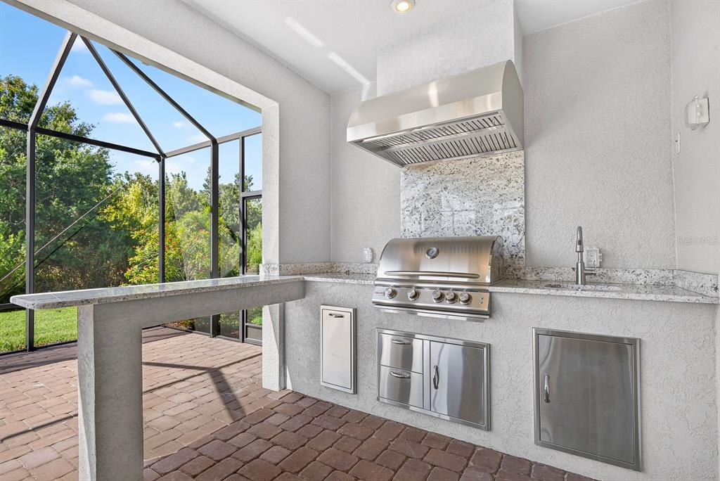An outdoor kitchen features a stainless steel grill, range hood, granite countertops, and brick flooring — 10924 WICKER PARK PLACE, PALMETTO, FL