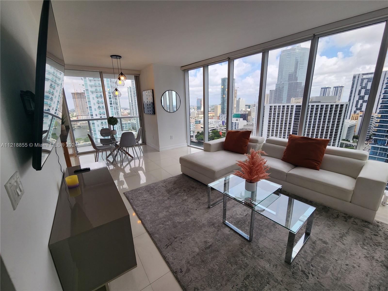 A modern living room features a white sectional sofa, glass coffee tables, and floor-to-ceiling windows — 485 Brickell Ave # 2504, Miami FL 33131, MIAMI, FL