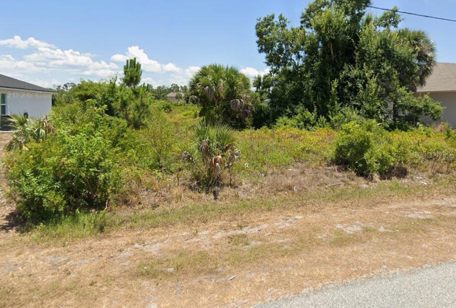 A vacant, overgrown residential lot sits between two existing houses under a clear blue sky — 9172 AGATE STREET, PORT CHARLOTTE, FL