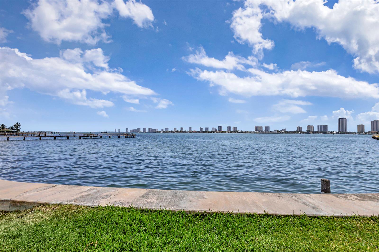 A concrete walkway borders a grassy lawn overlooking a bay with a distant city skyline — 2936 Lake Shore Drive # 309, Riviera Beach FL 33404, RIVIERA BEACH, FL