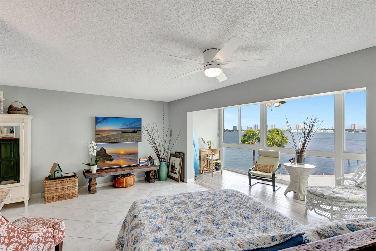A bedroom with a ceiling fan, two stacked televisions, and large windows overlooking the water — 2936 Lake Shore Drive # 309, Riviera Beach FL 33404, RIVIERA BEACH, FL