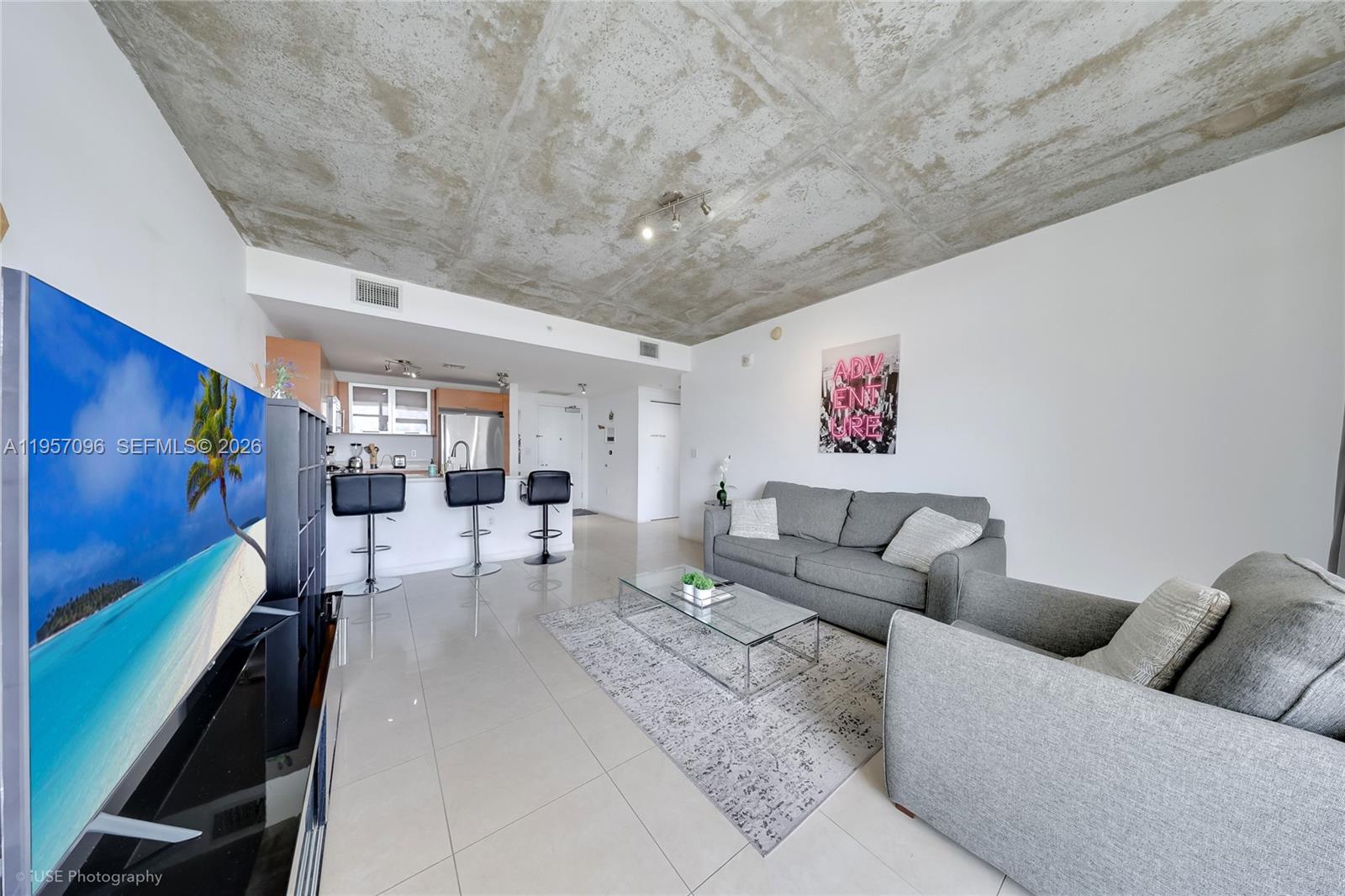 A living room features a concrete ceiling, tiled floors, a grey sofa, and an open kitchen — 3250 NE 1st Ave # 811, Miami FL 33137, MIAMI, FL