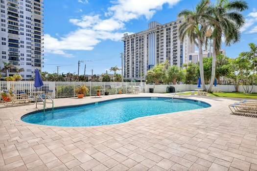 A kidney-shaped swimming pool sits on a paved patio surrounded by palm trees and buildings — 1901 S Ocean Drive # 202, Hollywood FL 33019, HOLLYWOOD, FL