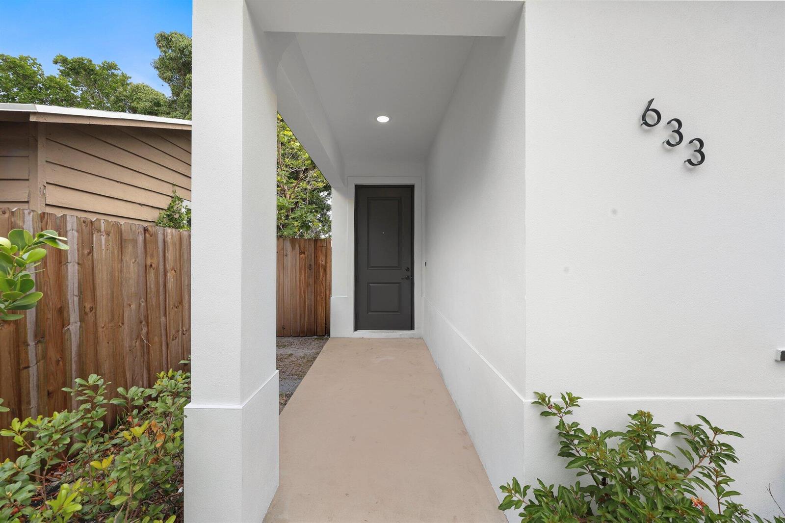 A covered entryway features a dark gray door, white stucco walls, and a concrete walkway — 633 55th Street, West Palm Beach FL 33407, WEST PALM BEACH, FL