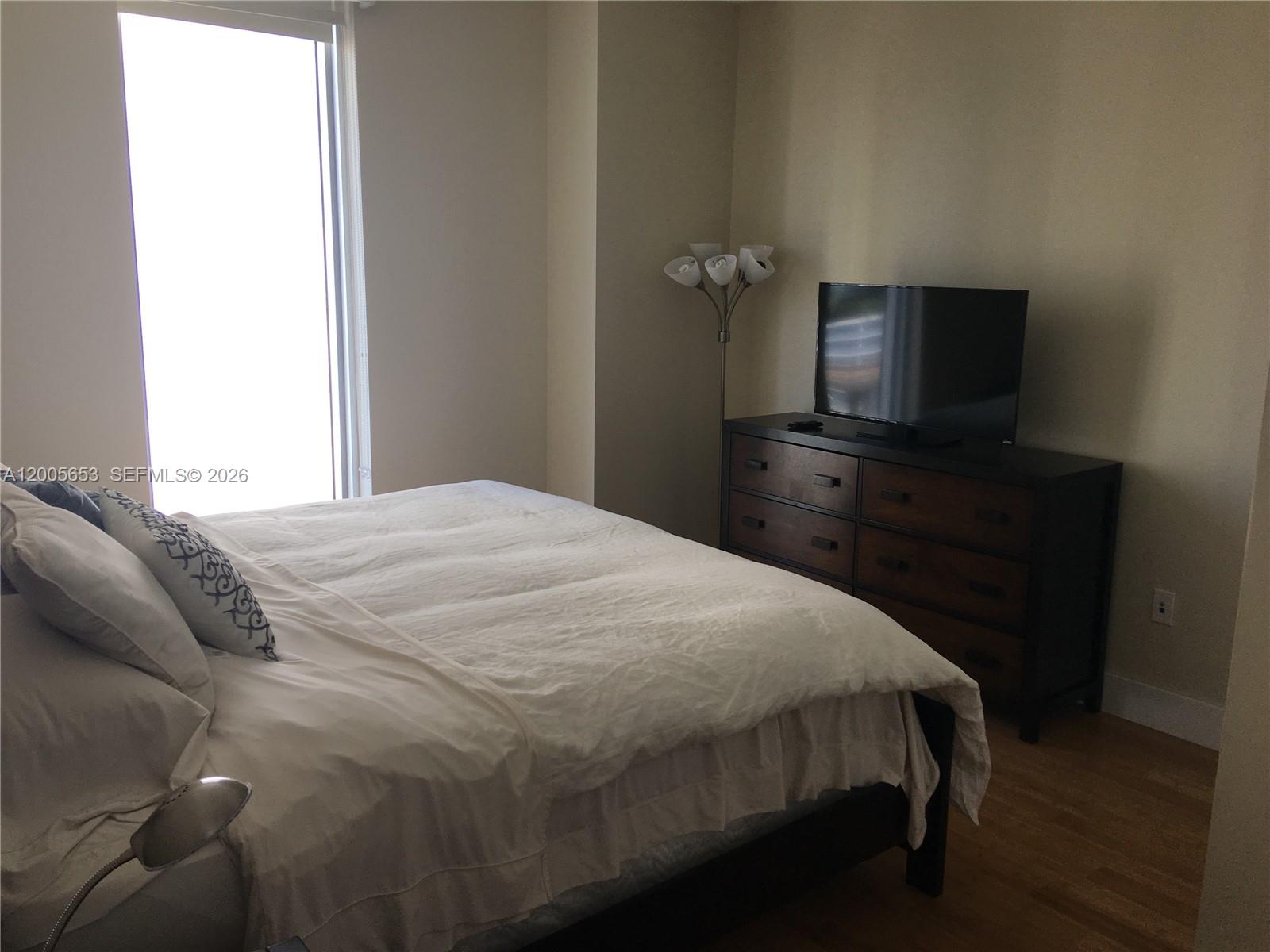 A bedroom features a bed with white bedding, a dark wood dresser, and a floor lamp — 185 SW 7th St # 1511, Miami FL 33130, MIAMI, FL