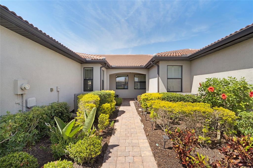 A paved walkway leads to the recessed front entrance of a stucco house with landscaping — 13908 ALAFAYA STREET, VENICE, FL