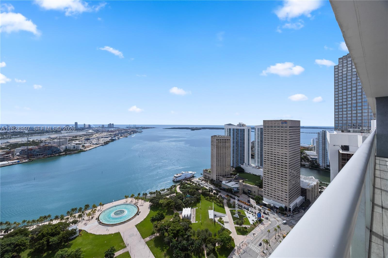 A high-angle view from a balcony overlooks a waterfront park, city buildings, and the ocean — 50 Biscayne Blvd # 4706, Miami FL 33132, MIAMI, FL