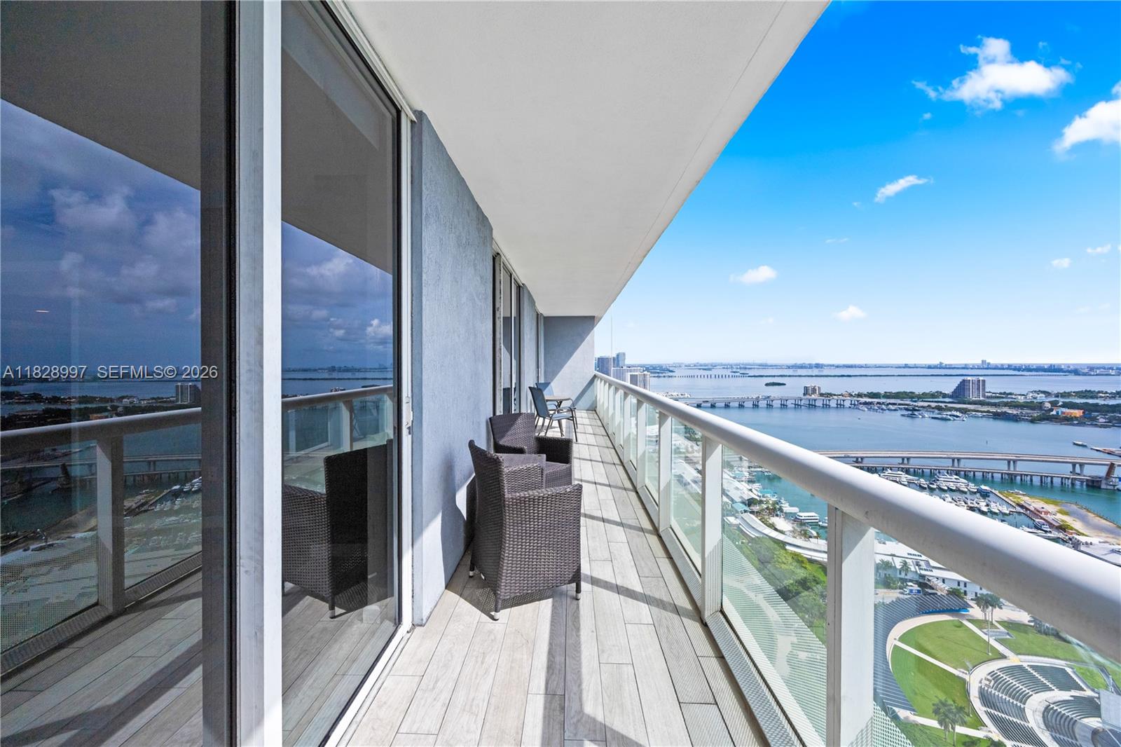 A high-rise balcony features wicker chairs, glass railings, and a panoramic view of the bay — 50 Biscayne Blvd # 4706, Miami FL 33132, MIAMI, FL
