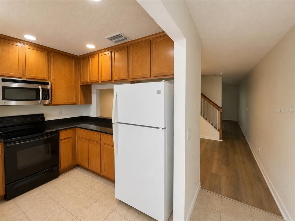 A kitchen with wood cabinets, black countertops, and a white refrigerator next to a hallway — 9410 CHARLESBERG DRIVE, TAMPA, FL
