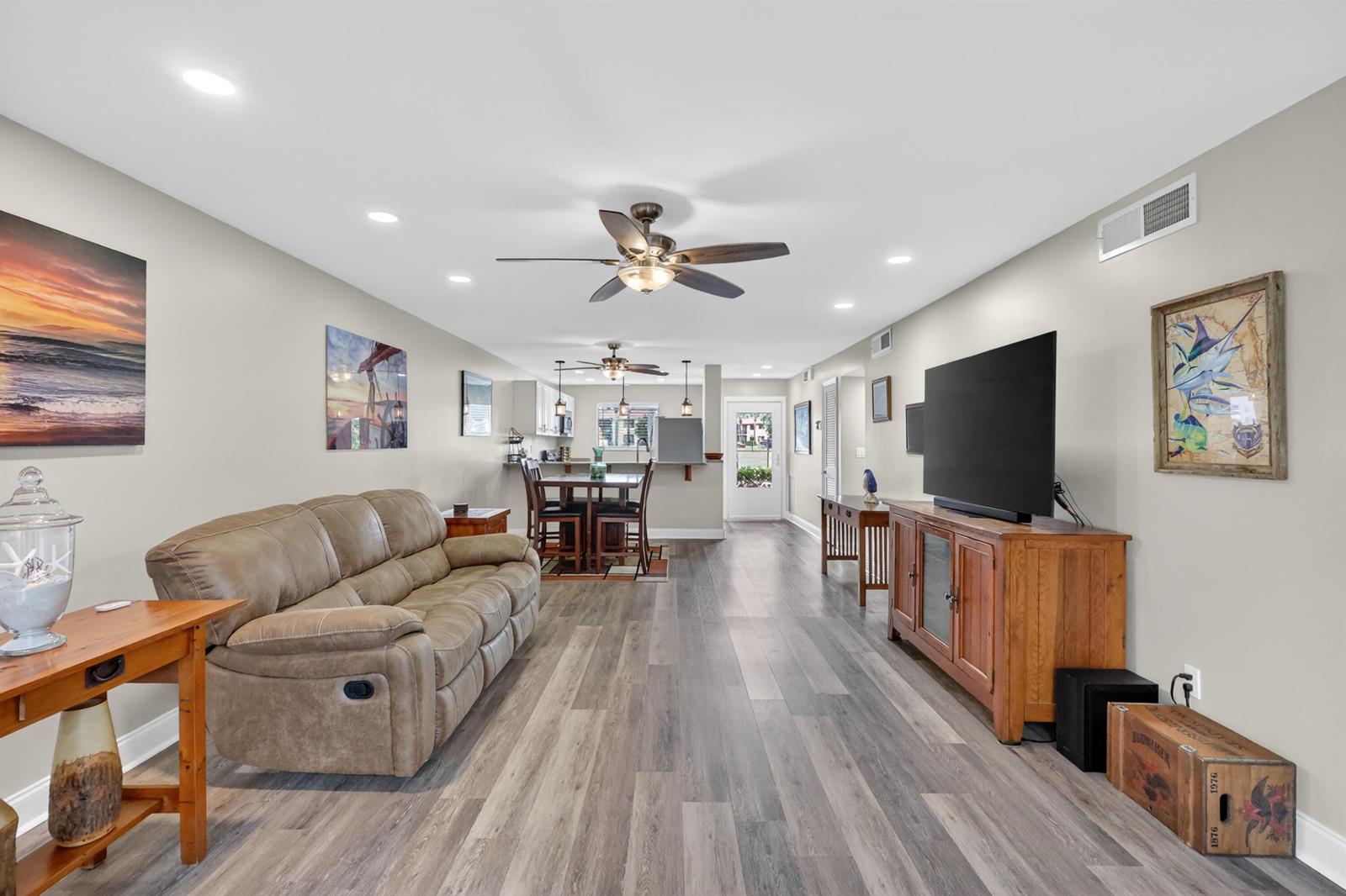 A living room with wood-look flooring, a brown sofa, a television console, and a kitchen — 1600 NE Dixie Highway # 104, Jensen Beach FL 34957, JENSEN BEACH, FL