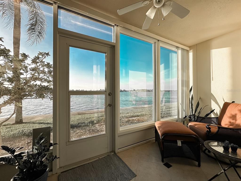 A sunroom features large windows overlooking a waterfront view, a ceiling fan, and patio furniture — 7920 SUN ISLAND DRIVE S UNIT 102, SOUTH PASADENA, FL