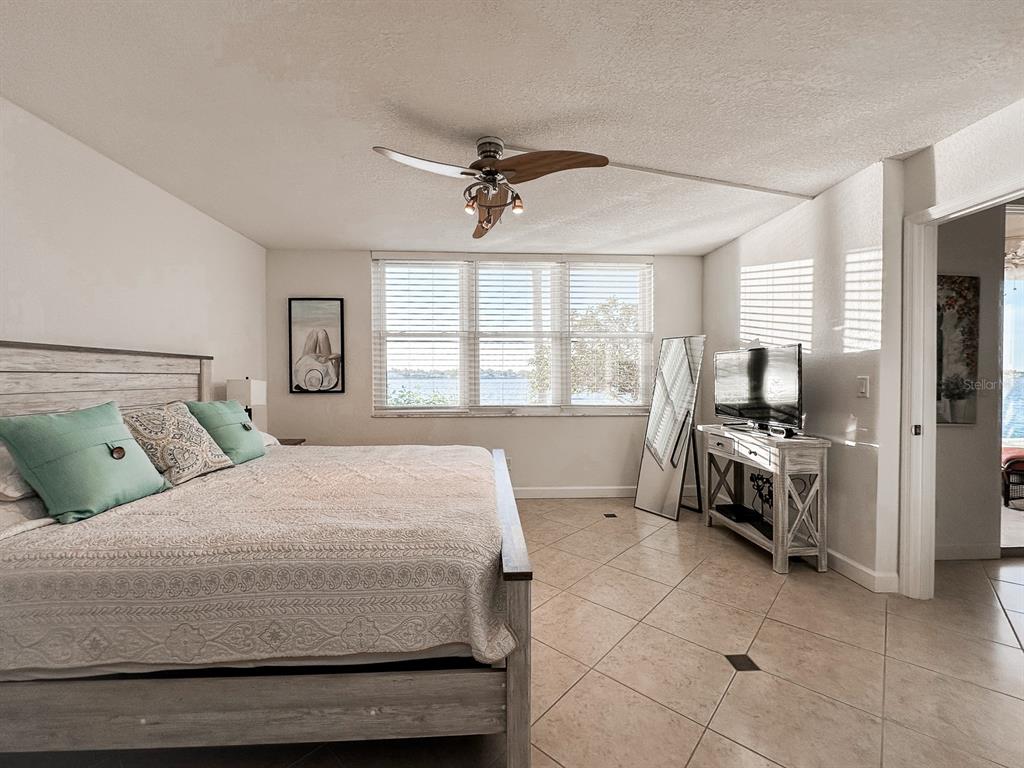 A bedroom features a king-sized bed, tile flooring, a ceiling fan, and a waterfront view — 7920 SUN ISLAND DRIVE S UNIT 102, SOUTH PASADENA, FL