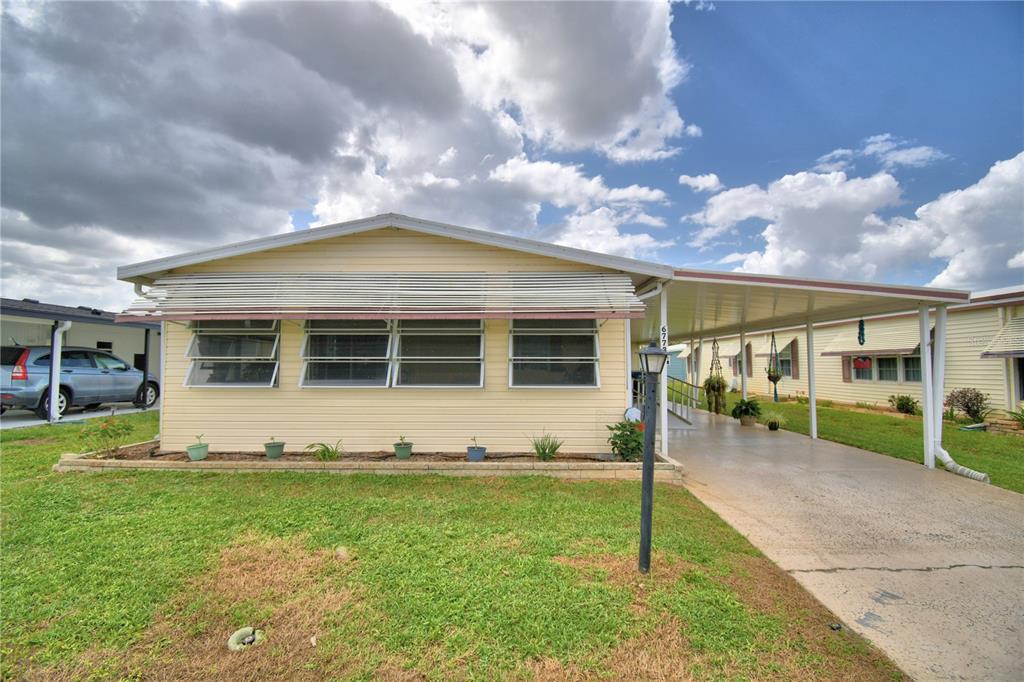 A light yellow manufactured home features a front carport, metal awnings, and a grassy lawn — 6773 BRENTWOOD DRIVE NE, WINTER HAVEN, FL