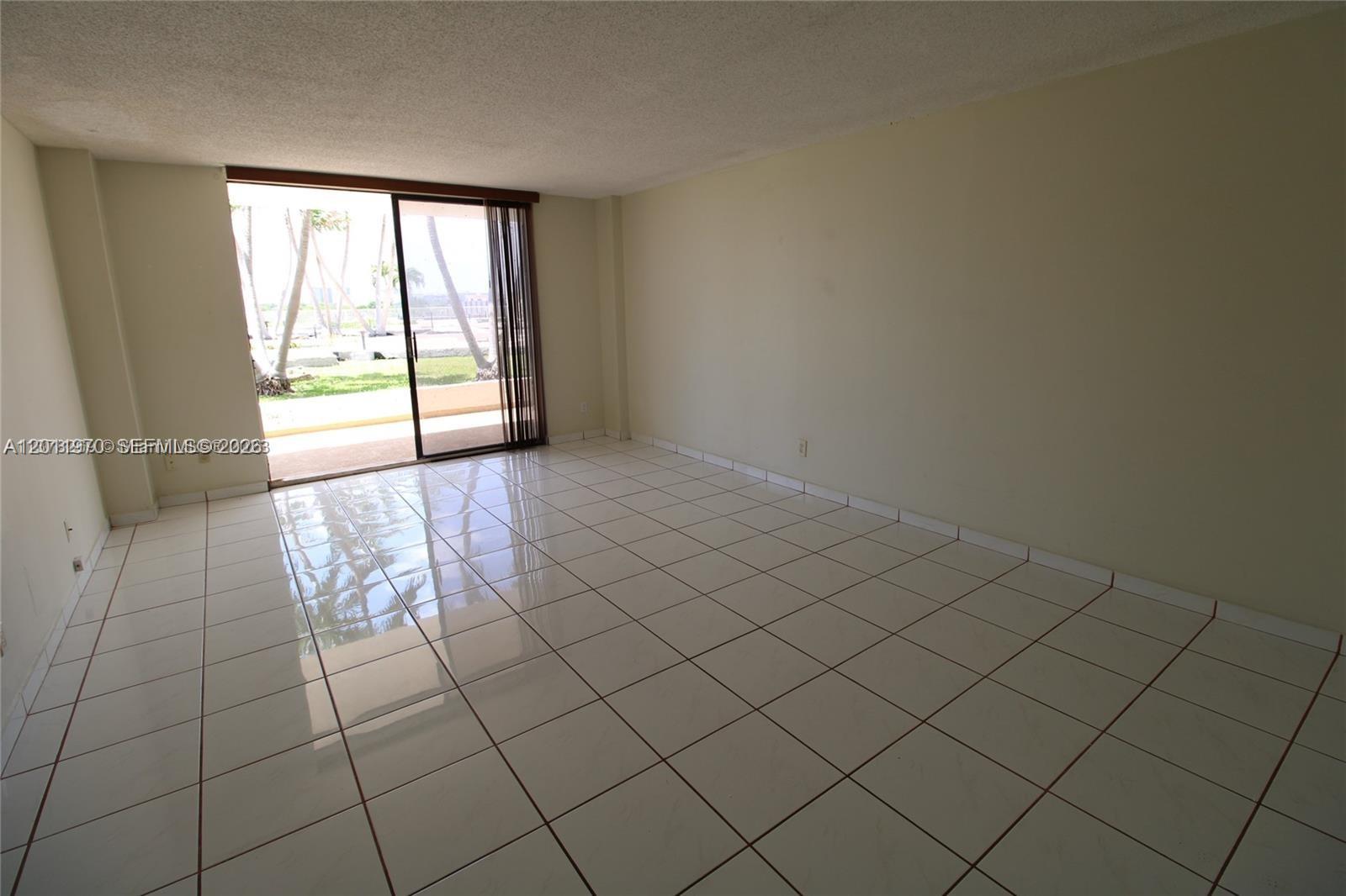 An empty room features glossy white floor tiles and a sliding glass door to a patio — 13499 Biscayne Blvd # 404, North Miami FL 33181, NORTH MIAMI, FL