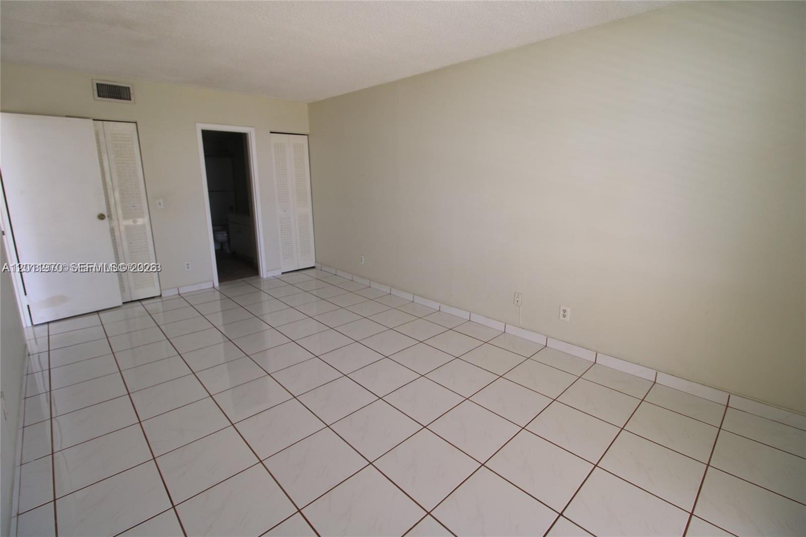 An empty room features white tiled floors, beige walls, and white louvered closet doors — 13499 Biscayne Blvd # 404, North Miami FL 33181, NORTH MIAMI, FL
