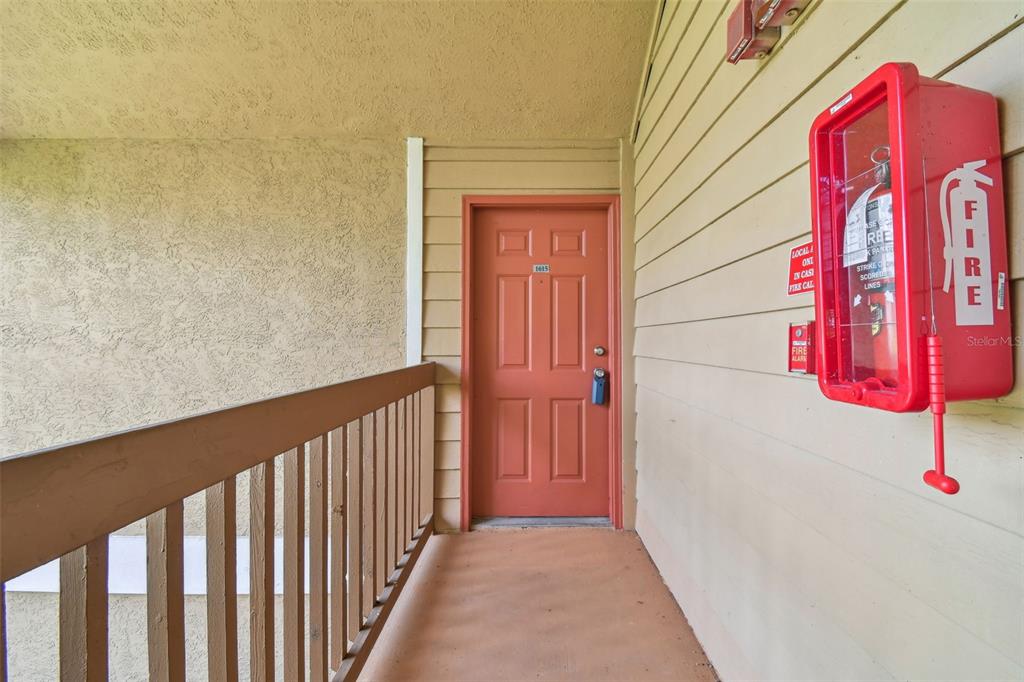 An exterior apartment hallway features a red door, a fire extinguisher cabinet, and a wooden railing — 9481 HIGHLAND OAK DRIVE UNIT 1615, TAMPA, FL