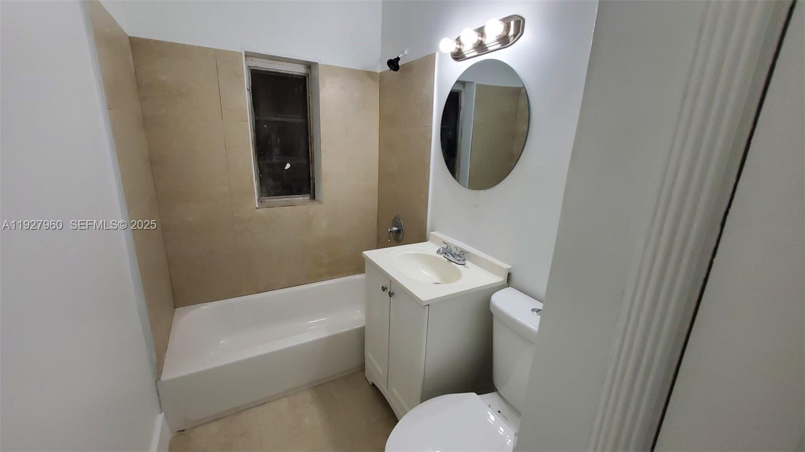 A small bathroom features a white vanity, a round mirror, and a bathtub with beige tiles — 560 NW 53rd St, Miami FL 33127, MIAMI, FL