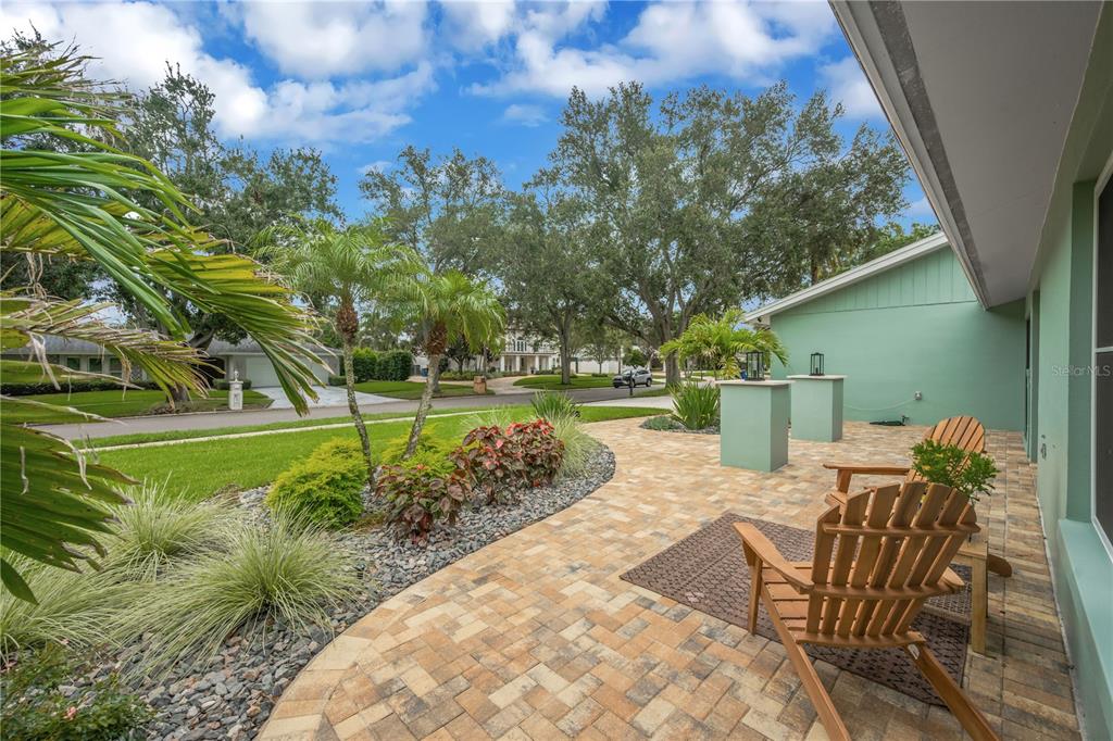 A paved patio with wooden chairs sits beside a green house exterior and landscaped yard — 2767 WESTCHESTER DRIVE N, CLEARWATER, FL