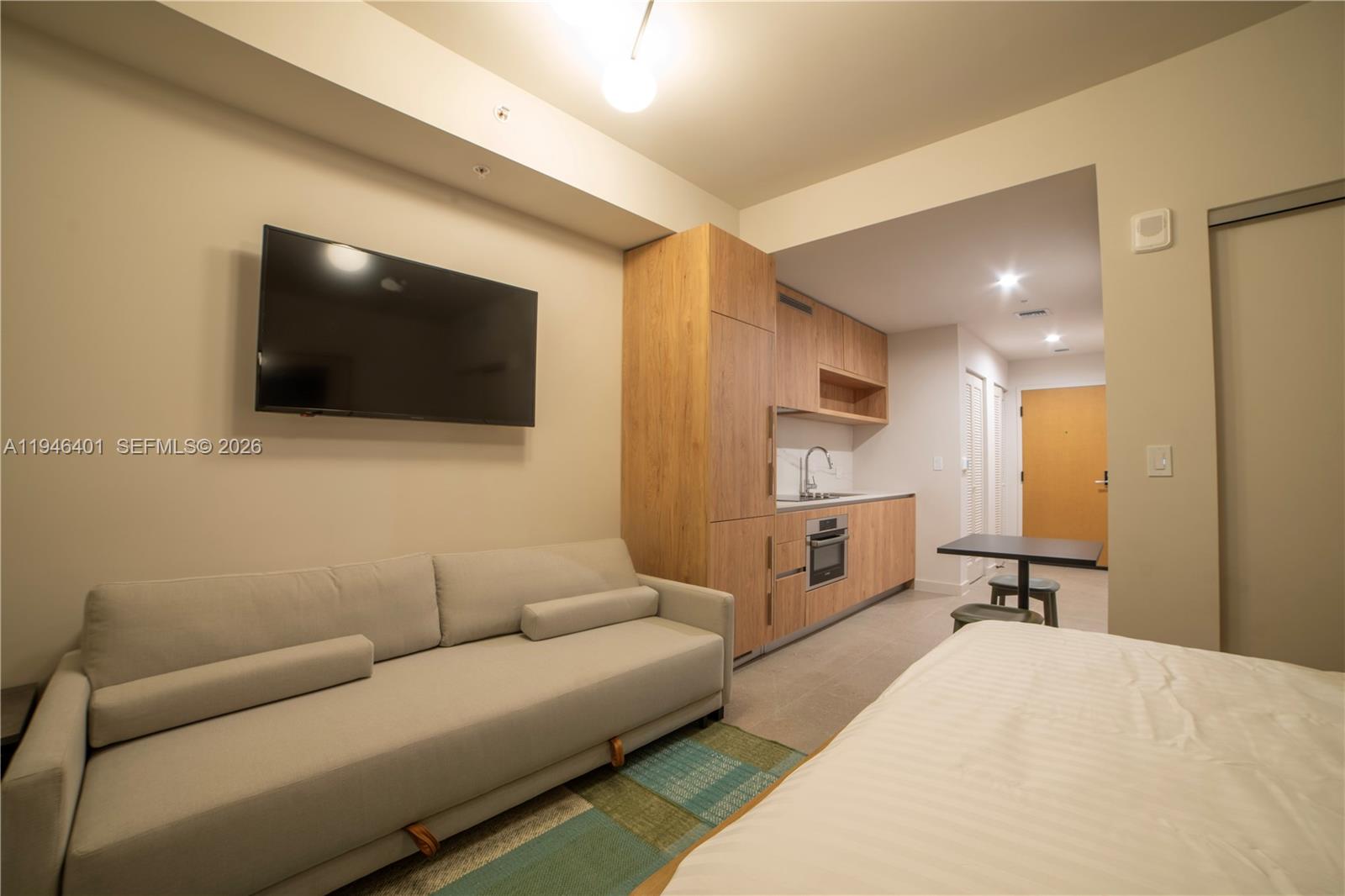 A studio apartment features a sofa, a wall-mounted television, and a kitchenette with wood cabinetry — 11 NE 6TH # 1908, Miami FL 33132, MIAMI, FL