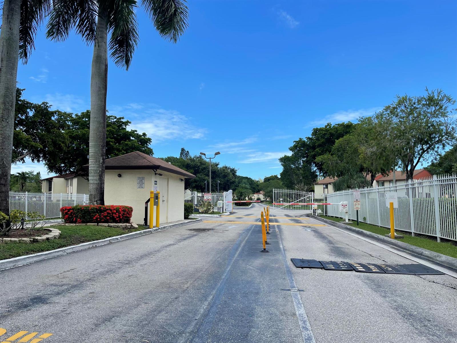 An entrance gate with a guardhouse, palm trees, and a white fence under a blue sky — 4857 Sable Pine Circle # B1, West Palm Beach FL 33417, WEST PALM BEACH, FL