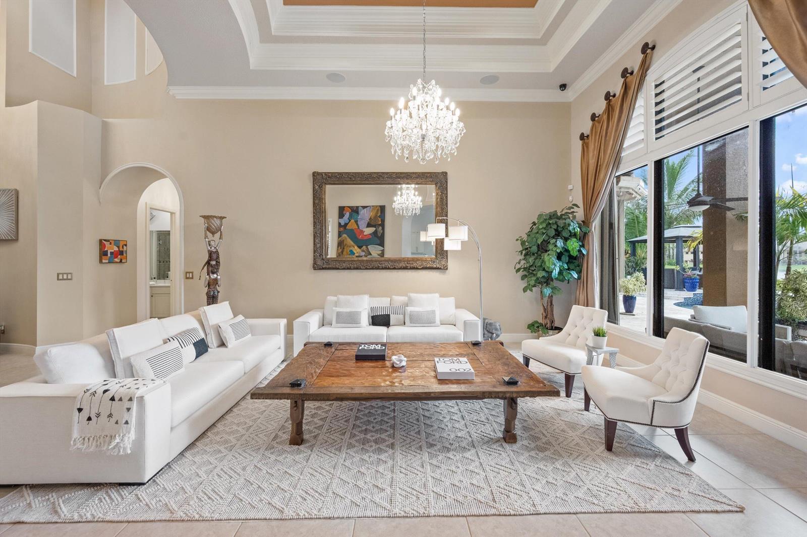 A bright living room features white sofas, a large wooden coffee table, and a crystal chandelier — 10771 Versailles Boulevard, Wellington FL 33449, WELLINGTON, FL